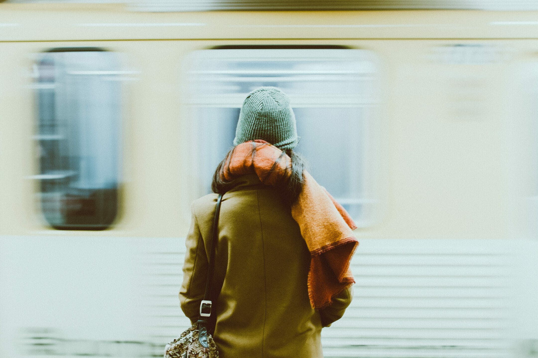 A woman is seen from behind as a subway train arrives in front of her. She is wearing winter clothes and carry a duffel bag.