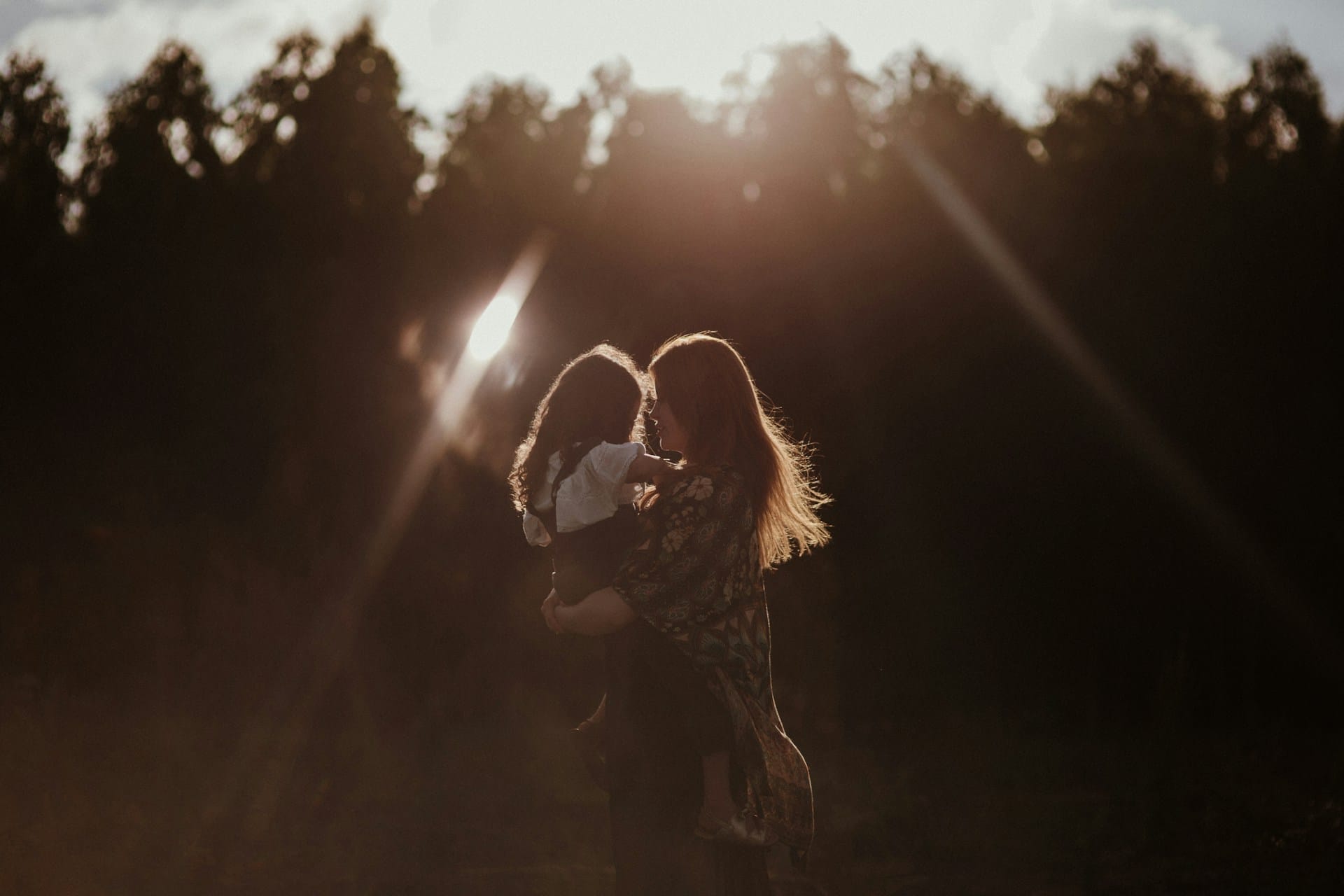 A woman holds up her small child as the sun goes down behind some trees, gently lighting the figures in a soft silhouette.